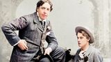Oskar Wilde im dunklen Anzug, stehend, neben ihm auf einer Bank Alfred Douglas mit Hut und weißen Hosen