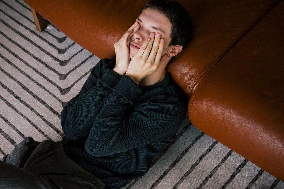 depressiver junger Mann sitzt mit den Händen vor dem Gesicht, ans Sofa gelehnt auf dem Teppich  1.5004