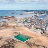 Strand mit befestigter Stadt im Hintergrund, davor ein Bassin voller Meerwasser, Luftaufnahme