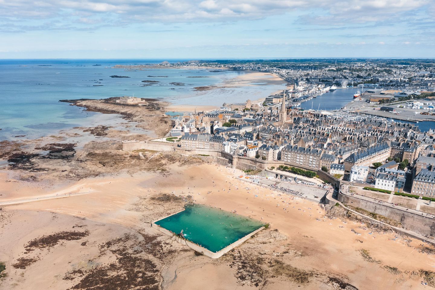 Strand mit befestigter Stadt im Hintergrund, davor ein Bassin voller Meerwasser, Luftaufnahme