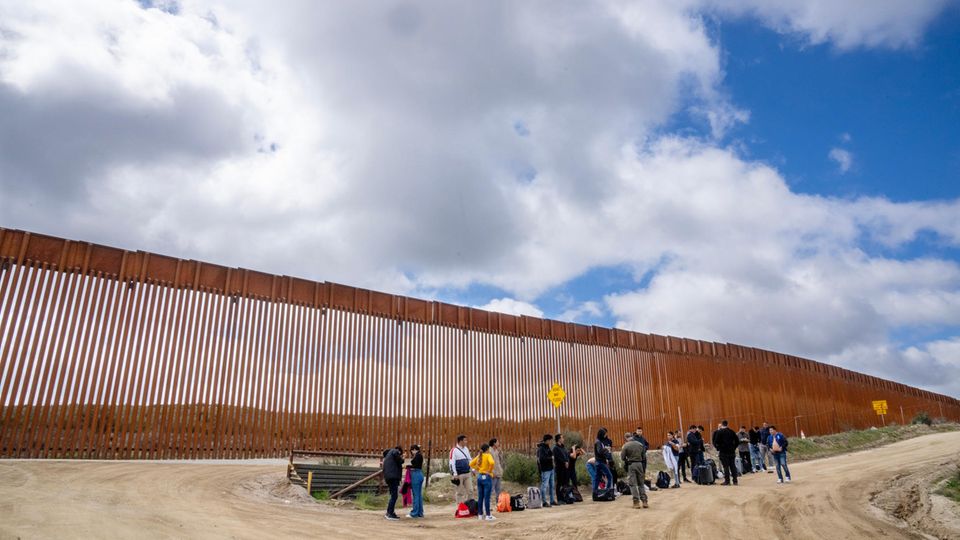 Menschen mit Gepäck stehen vor dem hohen Zaun zwischen Mexiko und den USA