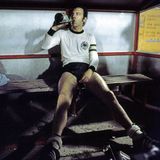 Franz Beckenbauer sitzt ausgelaugt auf einer Bank und trinkt aus einer Flasche