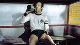 Franz Beckenbauer sitzt ausgelaugt auf einer Bank und trinkt aus einer Flasche