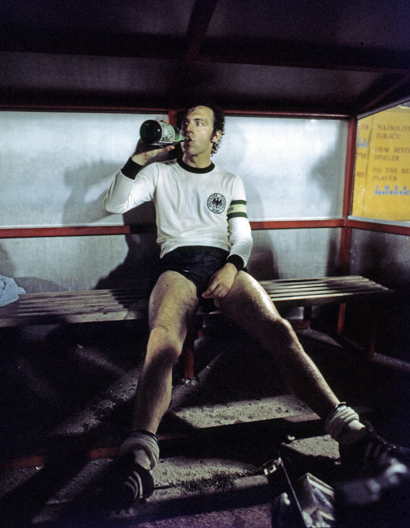 Franz Beckenbauer sitzt ausgelaugt auf einer Bank und trinkt aus einer Flasche