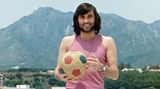 George Best posiert in kurzer Hose und Unterhemd mit einem Ball vor bergigem Hintergrund