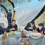 Ältere Menschen essen am Tisch mit Stäbchen und im Hintergrund steht ein Brautpaar mit Mikrophon