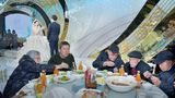 Ältere Menschen essen am Tisch mit Stäbchen und im Hintergrund steht ein Brautpaar mit Mikrophon