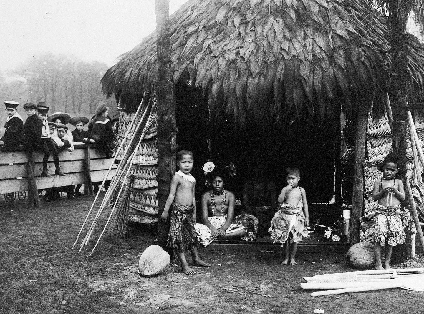 Kinder aus Samoa stehen vor einer traditionellen Hütte, beobachtet von einer Schülergruppe hinter einem Zaun