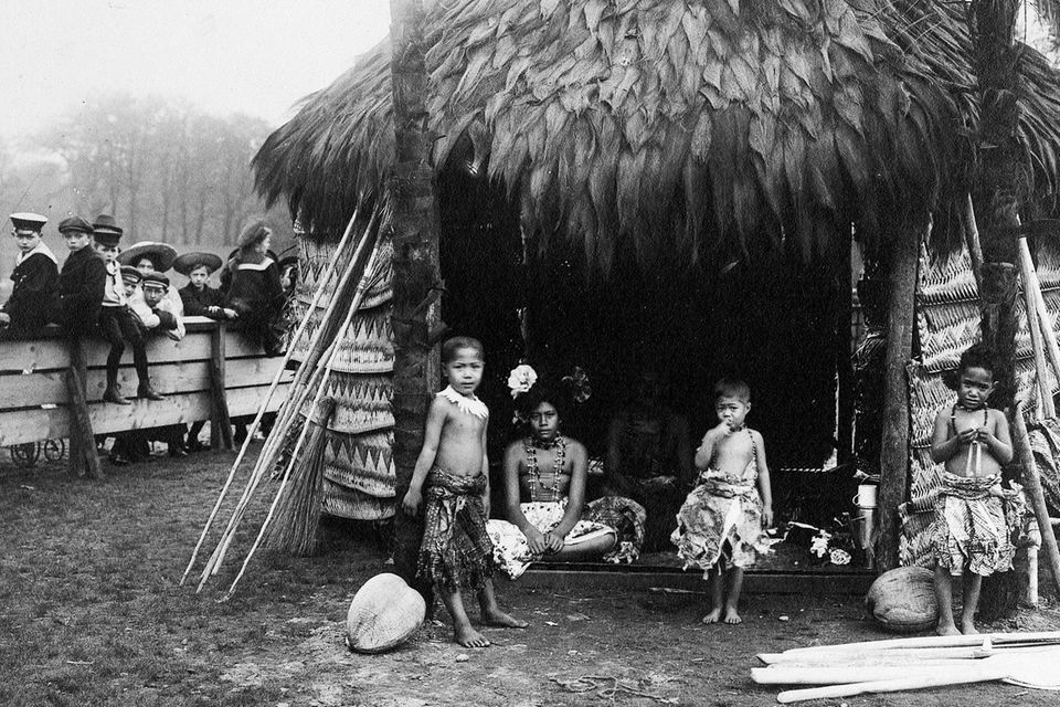 Kinder aus Samoa stehen vor einer traditionellen Hütte, beobachtet von einer Schülergruppe hinter einem Zaun 1.3492