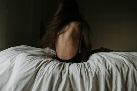 Eine dünne Frau sitzt mit dem Rücken zum Betrachter auf einem Bett