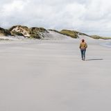 Frau geht am endlosen Strand, Dünen im Hintergrund,. Grauer Himmel.