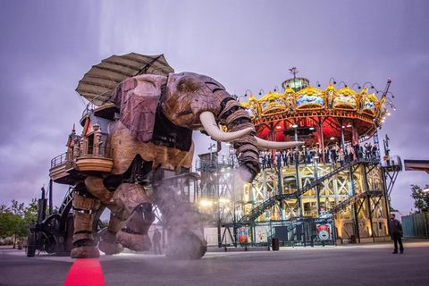 Beleuchtetes Karussell mit Meerestieren, auf dem Platz davor übergroßer künstlicher Elefant, Abendstimmung