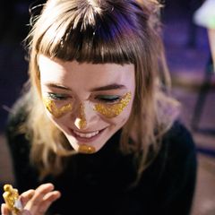 Eine junge Frau schminkt sich das Gesicht mit goldenem Glitter und lacht