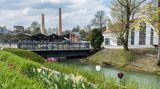 Schornstein, alte Gebäude, davor ein blühender Garten am Kanal