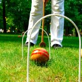 zwei Krocket-Tore stehen auf einem Rasen, dazwischen ein Ball, dahinter die Beine eines Spielers mit Schläger