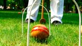 zwei Krocket-Tore stehen auf einem Rasen, dazwischen ein Ball, dahinter die Beine eines Spielers mit Schläger