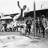 Sportler beim Hochsprung aus dem Stand, dahinter mehrere Schiedsrichter in Hut und Anzug