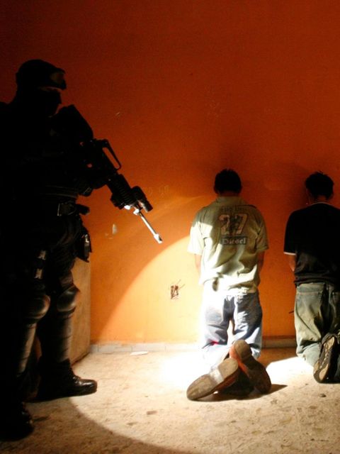 Gefangene knien vor einer Wand, dahinter ein bewaffneter Beamter