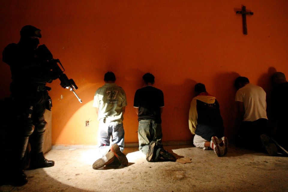 Gefangene knien vor einer Wand, dahinter ein bewaffneter Beamter 1.5468
