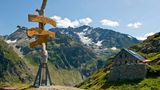 Blick auf eine Hütte auf der Windegg mit Wegweise im Vordergrund und Bergpanorama im Hintergrund.