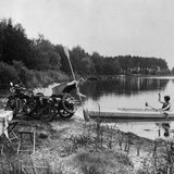 Ein Mann mit Motorrad und eine Frau im Faltboot am Ufer eines Sees