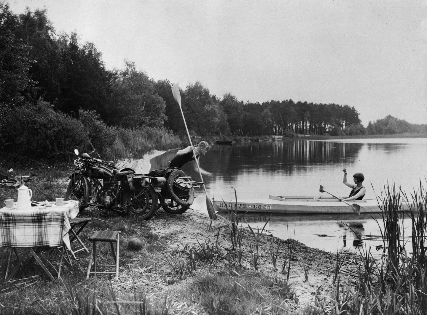 Ein Mann mit Motorrad und eine Frau im Faltboot am Ufer eines Sees