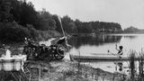 Ein Mann mit Motorrad und eine Frau im Faltboot am Ufer eines Sees