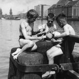 Drei Jungen sitzen auf Holzpfählen am Wasser und spielen Skat