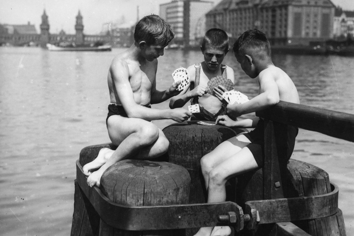 Drei Jungen sitzen auf Holzpfählen am Wasser und spielen Skat