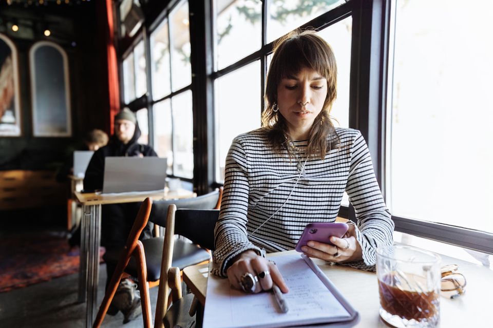 Junge Frau sitzt am Tisch  mit Unterlagen vor sich und starrt in ihr Handy Junge Frau sitzt am Tisch  mit Unterlagen vor sich und starrt in ihr Handy