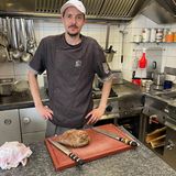 Mann in Kochhemd mit Cap vor Brett und zwei Messern in der Küche