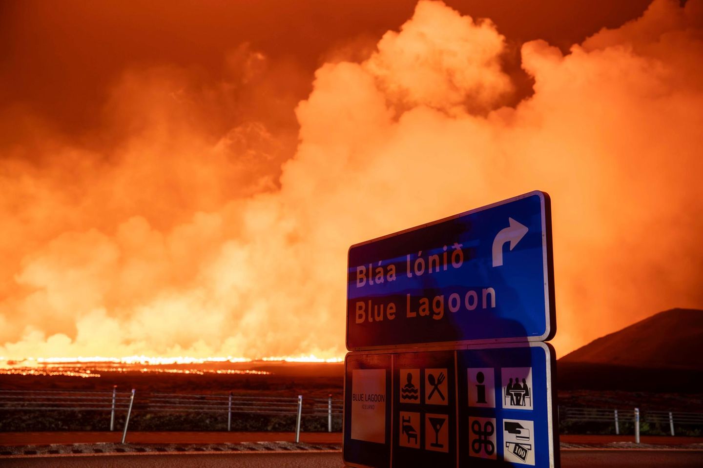 Schild mit dem Hinweis auf die blaue Lagune mit der neuen Eruption im Hintergrund am Vulkansystem Svartsengi