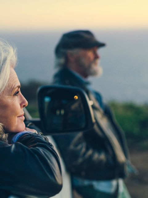 älteres Pärchen schaut entspannt in die Ferne,  der Mann verschwommen im Hintergrund  die Frau lehnt im Autofenster