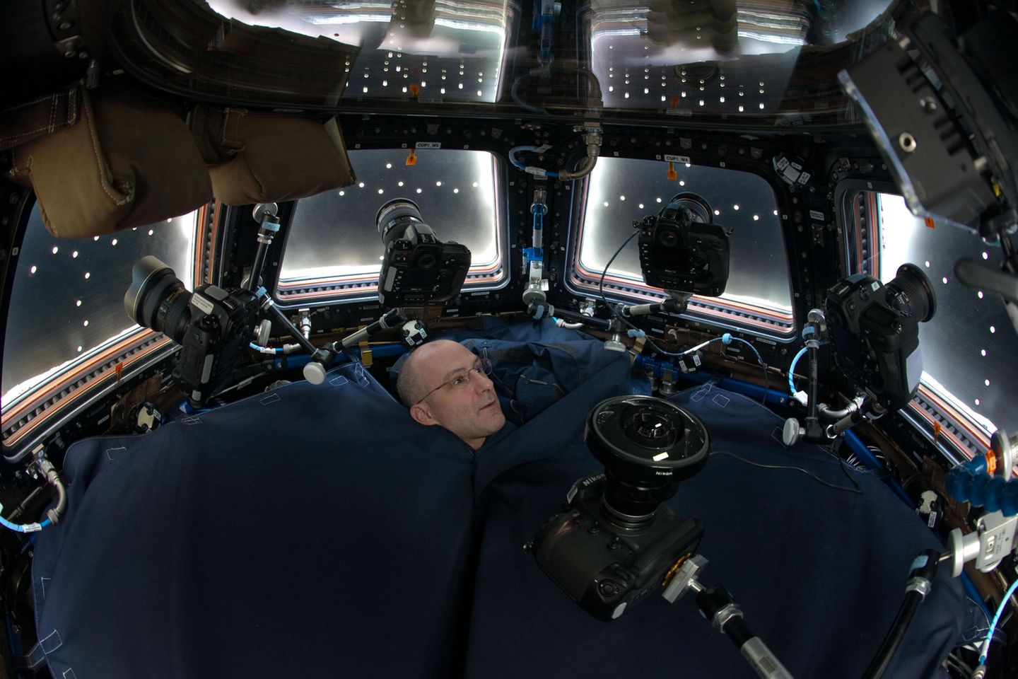 Trübe Aussichten Don Pettit 2012 inmitten der Spiegelreflex-Kameras, die er in der "Cupola" montiert hat. Die Aussichtskuppel des Moduls Tranquility ist meist der Erdseite zugewandt. Viel zu sehen gibt es im Moment dieser Aufnahme allerdings nicht: Spezielle Fensterläden verdunkeln den Raum. Sie dienen unter anderem dem Schutz der Scheiben vor Mikrometeoriten. Eine Stoffabdeckung (unten) schirmt die Kuppel vom Rest der Raumstation ab.