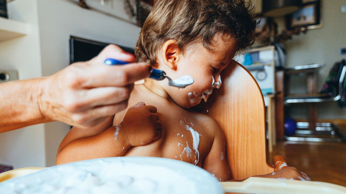 Ein beschmiertes Kleinkind wendet sich angewiedert von einem Löffel Joghurt ab