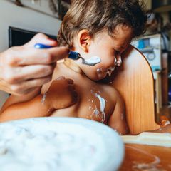 Ein beschmiertes Kleinkind wendet sich angewiedert von einem Löffel Joghurt ab