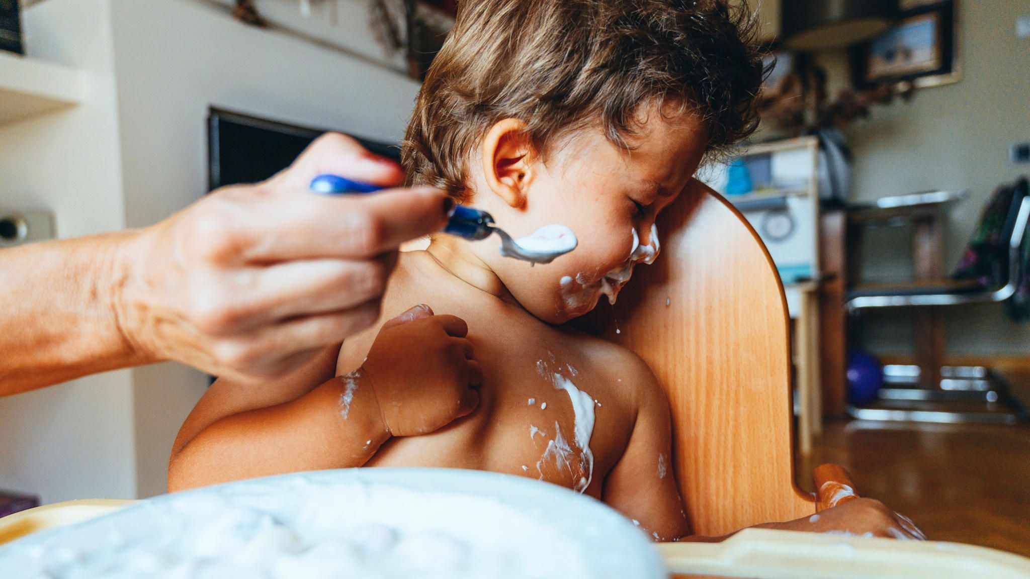 Ein beschmiertes Kleinkind wendet sich angewiedert von einem Löffel Joghurt ab