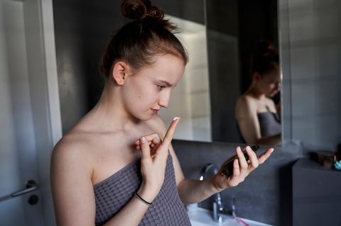Circa 12-jähriges Mädchen schaut in einen kleinen Handspiegel und führt Gesichtcreme mit dem Zeigefinger zum Gesicht.
