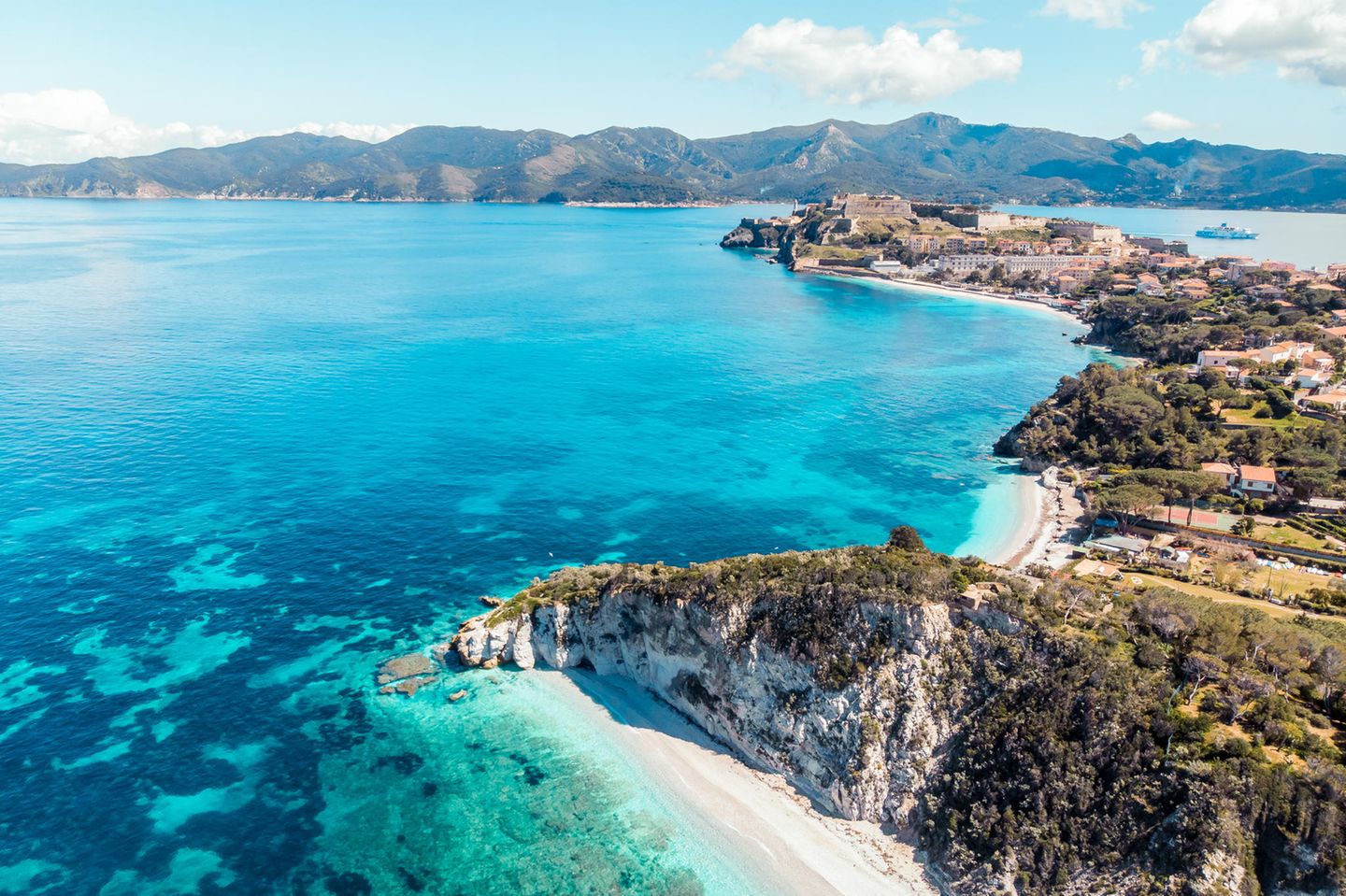 Strand mit Land und Häusern, dahinter sieht man Berge auf einer benachbarten Insel