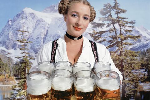 Eine Kellnerin auf dem Oktoberfest trägt acht Bierkrüge, im Hintergrund ein Alpenpanorama
