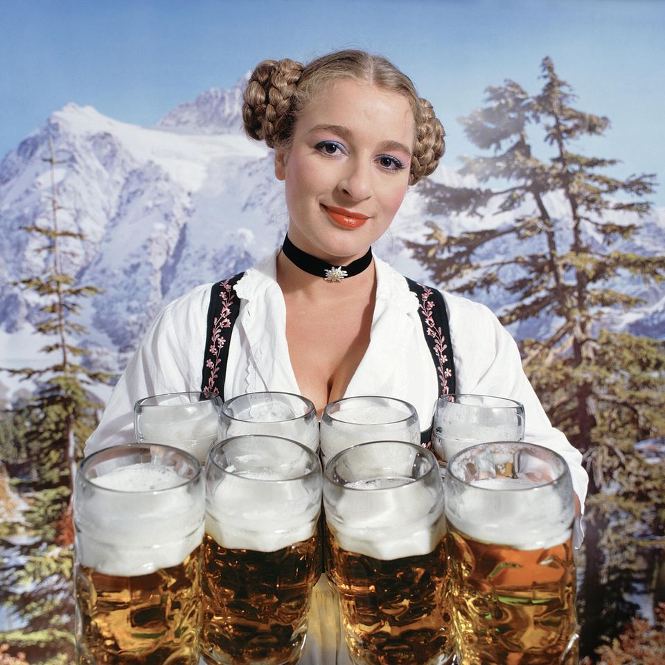 Eine Kellnerin auf dem Oktoberfest trägt acht Bierkrüge, im Hintergrund ein Alpenpanorama