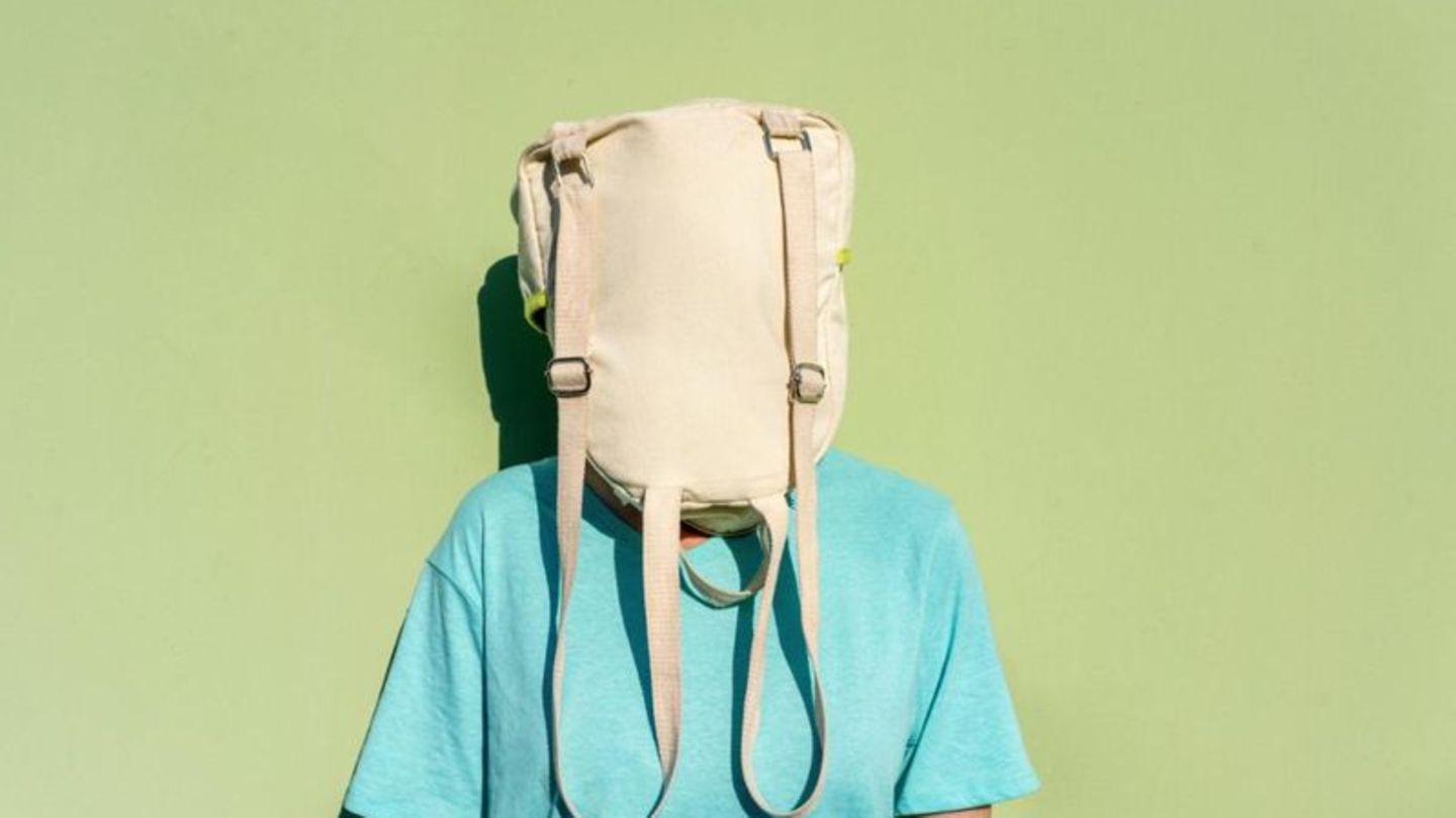 Eine Person in türkisem T-shirt steht vor einer grünen Wand und trägt einen Rucksack über dem Kopf