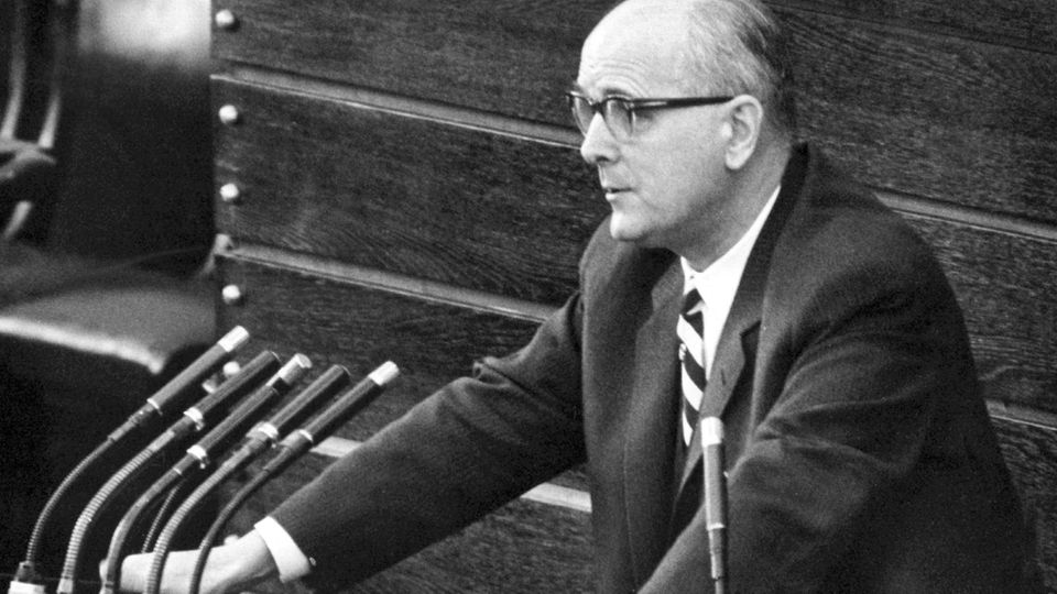Ein Mann mit Hornbrille und Krawatte steht an einem Rednerpult mit Mikrofonen Ein Mann mit Hornbrille und Krawatte steht an einem Rednerpult mit Mikrofonen