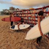 Bücherregale an einem Stadtstrand, in denen Surfer mit Surfbrettern unter dem Arm stöbern