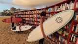 Bücherregale an einem Stadtstrand, in denen Surfer mit Surfbrettern unter dem Arm stöbern