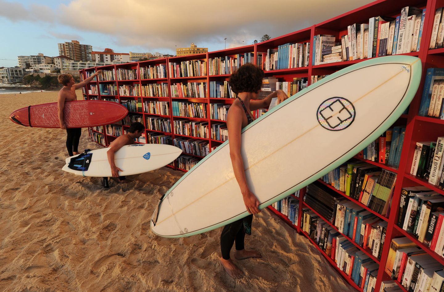 Billy on the Beach Bücherregale an einem Stadtstrand, in denen Surfer mit Surfbrettern unter dem Arm stöbern