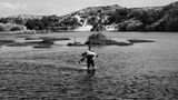 Schwarz-weiß Fotografie: Ein Mann trägt eine alte Frau durch einen Fluss