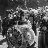 Schwarz-weiß Fotografie: Ein junges Mädchen hält Grabschmuck in der Hand. Hinter ihr die Trauernden mit Blumen