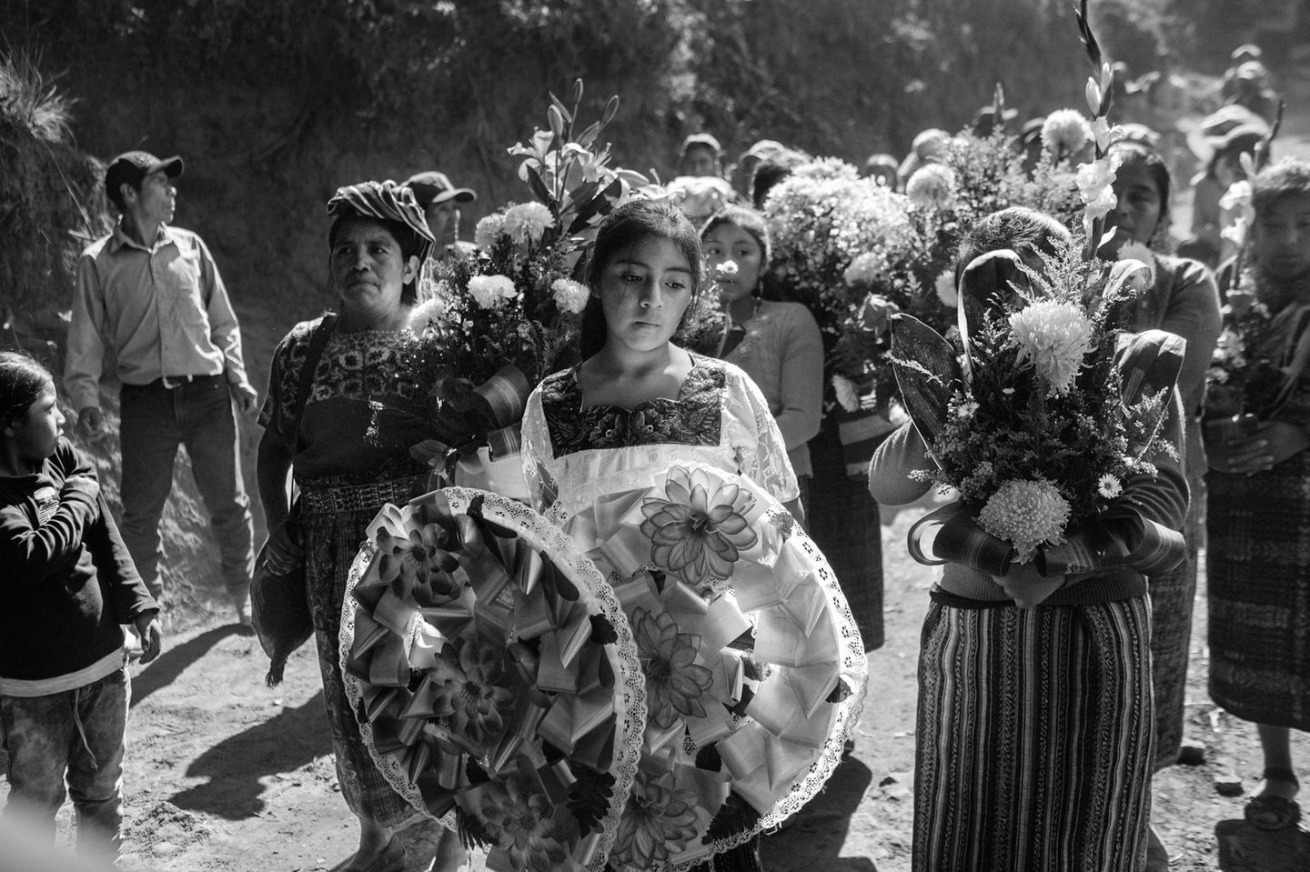 Guatemala Schwarz-weiß Fotografie: Ein junges Mädchen hält Grabschmuck in der Hand. Hinter ihr die Trauernden mit Blumen