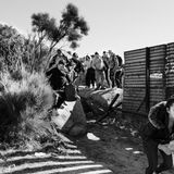 Schwarz-weiß Fotografie: Asylsuchende laufen durch eine Lücke im Grenzzaun zwischen Mexiko und USA